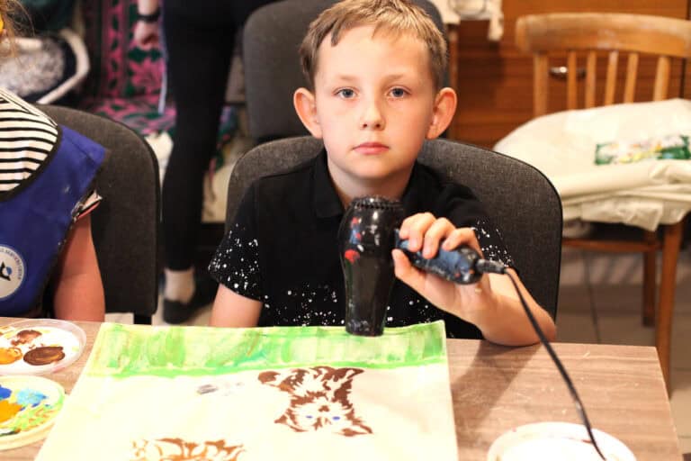 W centralnej części kadru chłopiec patrzący wprost do obiektywu. Siedzi na krześle, w dłoni trzyma suszarkę do włosów, którą suszy farbę na swojej pracy plastycznej