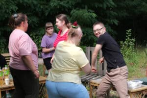 Na tle zieleni, pięć osób podczas tańca. Cztery dziewczyny i młodzieniec, który śmiejąc się patrzy wprost do obiektywu. Dziewczyny mają na sobie kolorowe, letnie koszulki, chłopiec czarną