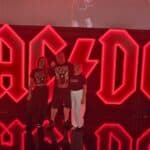 Łukasz Walczyński stoi w towarzystwie dwóch kobiet, wszyscy są ubrani w czarne koszulki z logo zespołu AC/DC. Za nimi czerwony, podświetlony neon z napisem „AC/DC”.