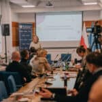 Na pierwszym planie siedzący przy stołach uczestnicy konferencji; w tle przy mównicy kobieta, za którą na ścianie wisi ekran multimedialny z wyświetlonym slajdem z tekstem "Spotkanie Ogólnopolskiej Sieci Turystyki Wytchnieniowej"