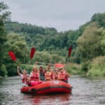 Siedmioosobowa grupa na czerwonym pontonie płynącym po wodzie; wszyscy mają na sobie kamizelki ratunkowe; a cztery osoby unoszą w górę wiosła; w tle widać zieleń drzew i krzewów