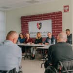 Na tle ścianki z napisem "Powiat Bolesławiecki" i herbem; przy długim stole siedzi sześć osób - cztery kobiety i dwóch mężczyzn; są zwróceni w kierunku dwóch mężczyzn na wózkach inwalidzkich