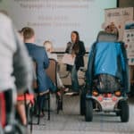 Kobieta z mikrofonem stoi na tle ekranu multimedialnego z napisem "Spotkanie Ogólnopolskiej Turystyki Wytchnieniowej 10.06.2025 r. Bukowina Tatrzańska"; widok z widowni