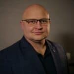 Łukasz Walczyński. Łysy mężczyzna w okularach patrzy prosto w obiektyw i delikatnie się uśmiecha. Ma na sobie ciemną koszulę i kraciastą, ciemną marynarkę. Zdjęcie jest portretem biznesowym, wykonanym na dość ciemnym tle, które podkreśla twarz i strój mężczyzny