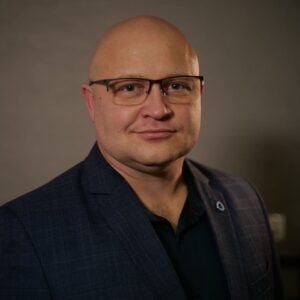 Łukasz Walczyński. Łysy mężczyzna w okularach patrzy prosto w obiektyw i delikatnie się uśmiecha. Ma na sobie ciemną koszulę i kraciastą, ciemną marynarkę. Zdjęcie jest portretem biznesowym, wykonanym na dość ciemnym tle, które podkreśla twarz i strój mężczyzny