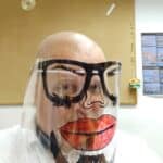 Mężczyzna w przezroczystej przyłbicy ochronnej ozdobionej karykaturalnym rysunkiem wykonanym flamastrami: ogromne czerwone usta, okulary z brwiami i nos