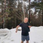 Uśmiechnięty mężczyzna stoi na śniegu w krótkich spodenkach i t-shircie, mimo zimowej scenerii. Z plecakiem na ramieniu i kciukiem uniesionym w górę pozuje na tle bezlistnego lasu.