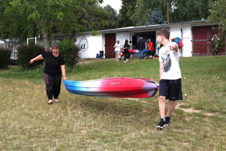 Na pierwszym planie dwie osoby: śmiejąca się młoda kobieta i młody mężczyzna, który na nią spogląda niosąc kajak w kolorze niebiesko czerwono białym. W tle budynek, przed którym stoi kilka osób