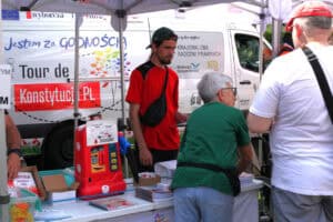 Stoisko Tour de Konstytucja i Krajowa Izba Radców Prawnych. Na tle białego busa z napisem Jestem za Godnością, Tour de Konstytucja stoi mężczyzna w czerwonej koszulce, ma na głowie czapkę typu „kaszkietówka” nałożoną daszkiem do tyłu. Przed stoiskiem, tyłem do obiektywu dwie osoby: kobieta i mężczyzna. Na stole przed nimi broszury i gadżety, wśród nich duża czerwona skarbonka
