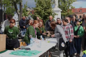 Kilkanaście osób. Od lewej: za podłużnym stołem (punkt zapisu uczestników) trzy kobiety w zielonych koszulkach Fundacji Pomoc Daje Moc. Na stole m. in.: koszulki fundacji, puszka na datki. Po przeciwnej stronie stołu osoby ustawione w kolejce, w tle budynki, zieleń drzew i błękit nieba