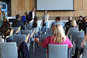 Sala wypełniona rzędami krzeseł, na których siedzą ludzie tyłem do obiektywu. Przed nimi stoi, od lewej: mężczyzna w okularach i kobieta. Za nimi ściana pokryta drewnianymi panelami, na niej widoczna jest dolna część ekranu