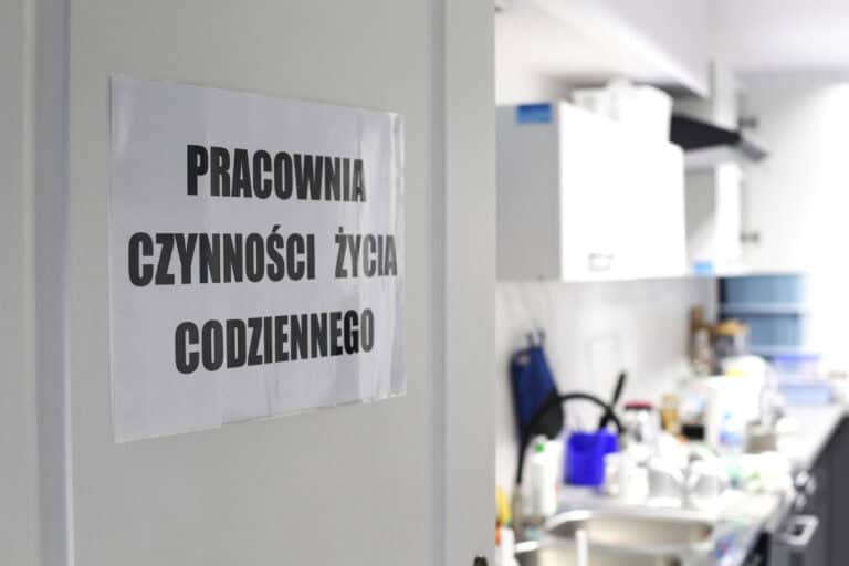 Od lewej: na uchylonych w głąb pracowni białych drzwiach kartka z napisem: Pracownia Czynności Życia Codziennego. Z prawej: nieostry widok na wnętrze pracowni