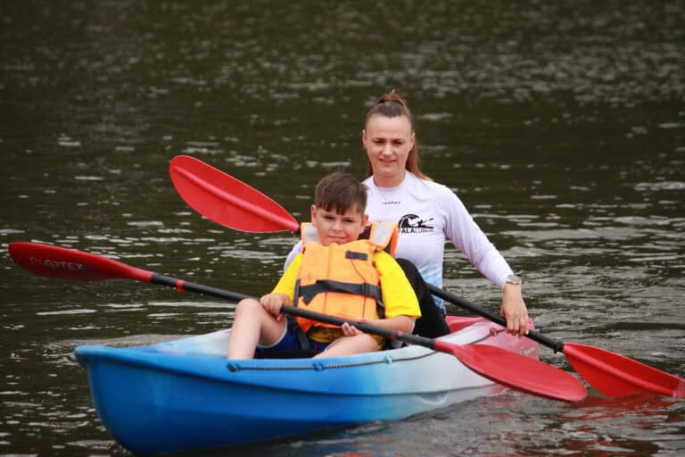 Niebieski kajak na wodzie, a w nim chłopiec w pomarańczowym kapoku, za nim instruktorka – młoda kobieta. Oboje trzymają czerwone wiosła. Otacza ich woda