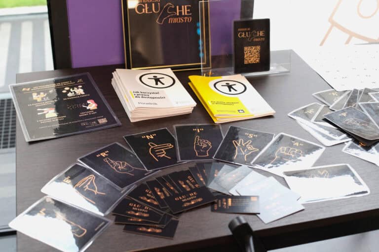 Na czarnym stoliku leżą poradniki o dostępności i małe czarne obrazki uczące jak komunikują się osoby z niepełnosprawnością słuchu. Są one ułożone na kształt półkola. Na skraju stolika stoi wydruk w ramkach z napisem Fundacja Głuche Miasto