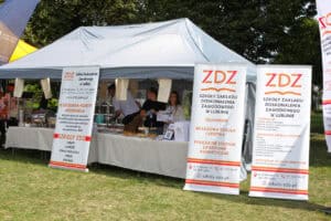 Biały namiot, przed którym stoją trzy banery ZDZ, Szkoły Zakładu Doskonalenia Zawodowego w Lublinie. W środku namiotu kilka osób z obsługi, w tle zieleń drzew
