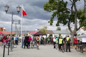 Od lewej linia latarni i maszt z biało-czerwoną flagą. Dolna część kadru to grupy osób stojące przy rowerach. Górną połowę kadru wypełnia zachmurzone niebiesko-szare niebo. Po prawej stare drzewo z rozłożystą koroną
