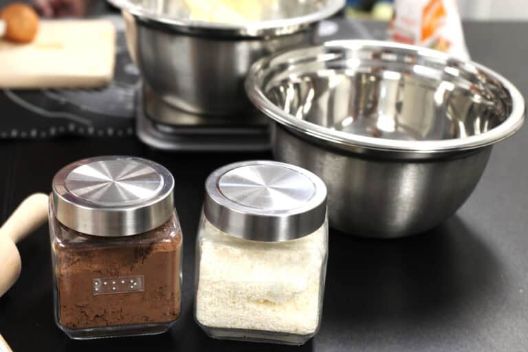 Na pierwszym planie dwa szklane pojemniki z metalowymi nakrętkami. W pojemniku od lewej kakao, w drugim wiórki kokosowe. Na pojemnikach przezroczyste etykiety z nazwami produktów napisane alfabetem Braille’a. W tle metalowe miski kuchenne