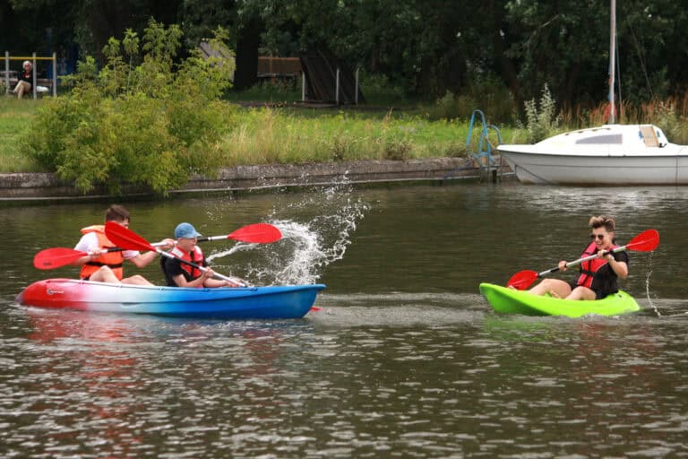 Na pierwszym planie dwa kajaki. Od lewej czerwono biało niebieski, a w nim dwaj młodzieńcy wiosłami rozchlapują wodę. Rozbryzgi wody kierują w stronę drugiego kajaka, w którym siedzi młoda kobieta w kapoku. Kobieta ma okulary przeciwsłoneczne, trzyma wiosło, śmieje się