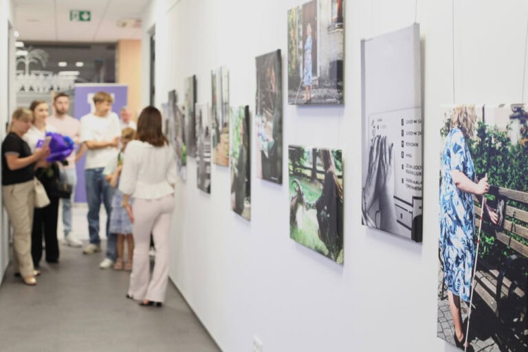 Fotografia w perspektywie zbieżnej. Ostrość z prawej strony kadru na zdjęcia wiszące na jasnej ścianie. W głębi z lewej grupa kilku osób