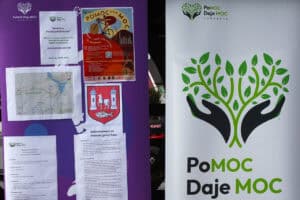 Od lewej plakaty Pomoc Daje Moc, mapa z trasą przejazdu, plakat z herbem Kocka. Z prawej plakat Fundacji Pomoc Daje Moc i logo w postaci rozłożonych dłoni, z których wyrasta drzewo