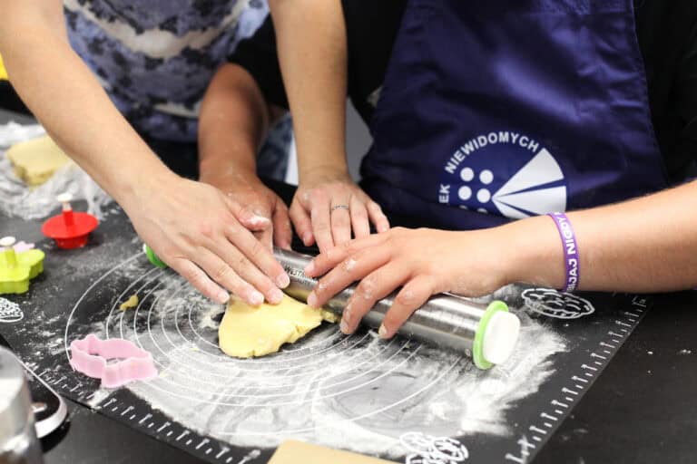 W centralnej części zbliżenie na ręce oprószone mąką, wałkujące kawałek ciasta na czarnej macie kuchennej. W lewym dolnym rogu trzy foremki do ciastek: różowa, czerwona i pistacjowa
