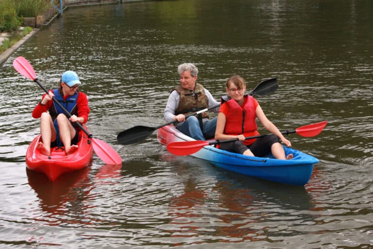 Na pierwszym planie dwa kajaki. Od lewej czerwony, a w nim młody człowiek. Kajak z prawej jest czerwono biało niebieski, siedzą w nim dwie osoby: młoda kobieta i mężczyzna. Oboje trzymają wiosła. Otacza ich woda