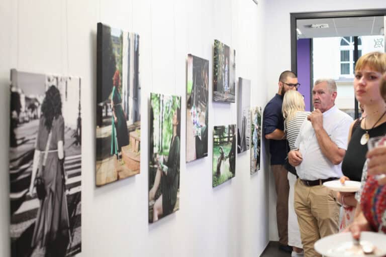 Perspektywa zbieżna. Od lewej fotografie wiszące na jasnej ścianie. W głębi korytarza grupa osób pogrążonych w rozmowie