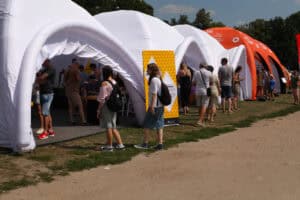 Ogólny widok na ciąg białych i pomarańczowych namiotów, przed którymi i w środku których znajdują się grupy ludzi. Na planie pierwszym droga utwardzona z pasem trawy, w tle błękit nieba i zieleń drzew