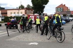 Na pierwszym planie rowerzyści w jaskrawozielonych kamizelkach wyjeżdżają z placu na ulicę. Na ich czele pojazd pilotujący i wóz strażacki. W tle zabudowania, lekko zachmurzone niebo
