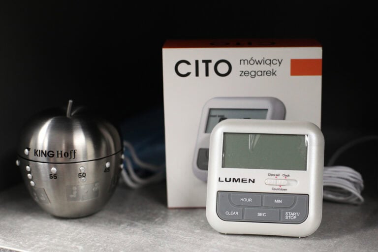 Od lewej minutnik kuchenny ze stali nierdzewnej. Po prawej wyjęty z opakowania stojącego z tyłu, biały kwadratowy zegarek mówiący