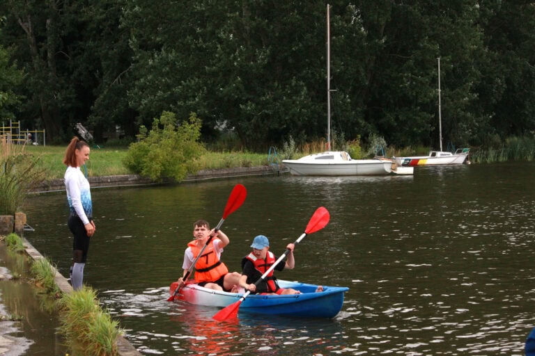 Na pierwszym planie od lewej na brzegu stoi młoda kobieta – instruktorka. Spogląda na wodę, na której w niebiesko biało czerwonym kajaku siedzą dwaj młodzieńcy. Obydwaj trzymają wiosła. W tle z prawej dwie białe łodzie tuż przy brzegu. Za nimi ciemna zieleń drzew