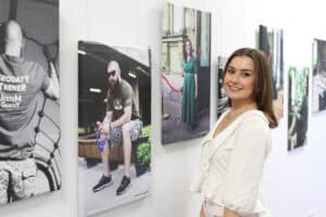 Fotografia pamiątkowa. Autorka prezentowanych zdjęć - Paulina Muszyńska pozuje na tle jasnej ściany, na której wiszą asymetrycznie fotografie. Kobieta stoi bokiem, uśmiecha się do obiektywu. Ma na sobie jasną bluzkę z dużym ozdobnym kołnierzykiem