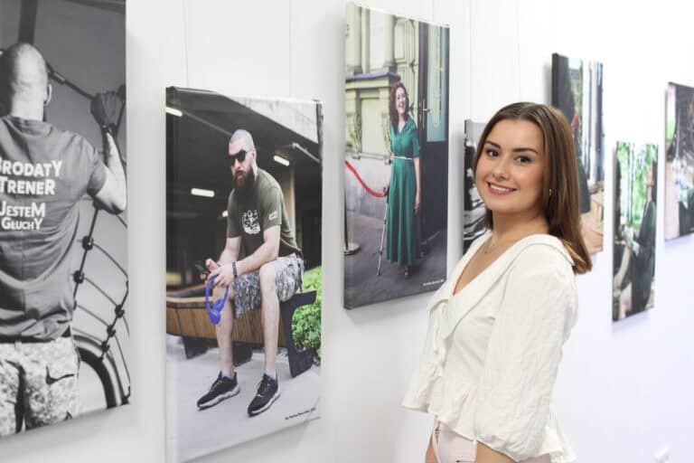 Fotografia pamiątkowa. Autorka prezentowanych zdjęć - Paulina Muszyńska pozuje na tle jasnej ściany, na której wiszą asymetrycznie fotografie. Kobieta stoi bokiem, uśmiecha się do obiektywu. Ma na sobie jasną bluzkę z dużym ozdobnym kołnierzykiem