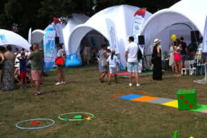 Na pierwszym planie, na trawie porozkładane kolorowe plansze i koła typu hula hop, a także duża zielona kostka do gry. W dalszej perspektywie grupki osób, ustawione przed białymi namiotami
