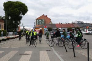 Rowerzyści wyjeżdżający z placu na drogę. Niektórzy mają na sobie jaskrawozielone kamizelki. Otoczenie to miejskie zabudowania, niebo niebiesko-szare, zachmurzone