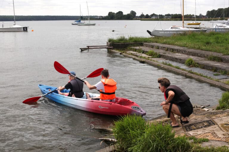 Na pierwszym planie zwodowany kajak z dwoma młodymi mężczyznami w kapokach. Z prawej, na brzegu kobieta spogląda na kajak. W tle woda i kilka żaglówek