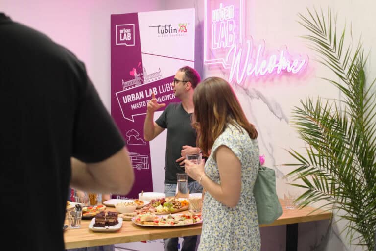 Przy stole z przekąskami dwie osoby. Kobieta stojąca tyłem, w ręku trzyma szklankę. Mężczyzna po drugiej stronie stołu gestykuluje patrząc przez ramię. W tle stoi baner "Urban Lab". Na ścianie z prawej różowy neon Urban Lab Welcome