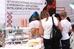 Stoisko z napisem: Wykorzystanie rozwiązań cyfrowych i sztucznej inteligencji w medycynie – projekt badawczy". Przy stoisku z prawej grupa osób. Od lewej fragment ekspozycji: kolorowe modele okulistyczne i gablota z okularami