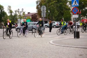Kilka osób na rowerach. Kobieta prowadząca grupę wykonuje gest lewą ręką sygnalizujący zakręt. W tle miejskie zabudowania