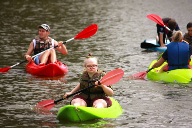 Kilka osób w kajakach. Na pierwszym planie w jaskrawo zielonym kajaku płynie młoda kobieta w okularach. Trzyma wiosło, które jedną stroną zanurzone jest w wodzie. Za nią, na drugim planie z lewej młody mężczyzna płynie w czerwonym kajaku