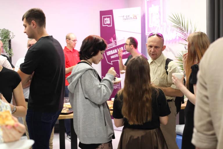 Na tle baneru Urban Lab Lublin stoi kilka osób pogrążonych w rozmowie. Kobieta w centrum kadru ma przed sobą talerzyk, inna trzyma w ręku kubek. Dwie osoby stoją tyłem do obiektywu
