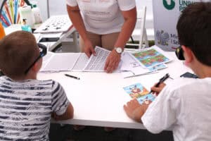 Przy stoliku, tyłem do obiektywu dwaj chłopcy. Przed nimi porozkładane kartki z obrazkami, napisami. Obaj mają na sobie specjalistyczne okulary. W centrum kadru ręce kobiety na małym stosie kartek