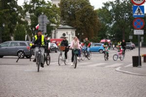W centralnej części kadru grupa rowerzystów, wśród nich dzieci na mniejszych rowerkach. Uchwyceni na zakręcie drogi. W tle miejskie zabudowania i zieleń drzew