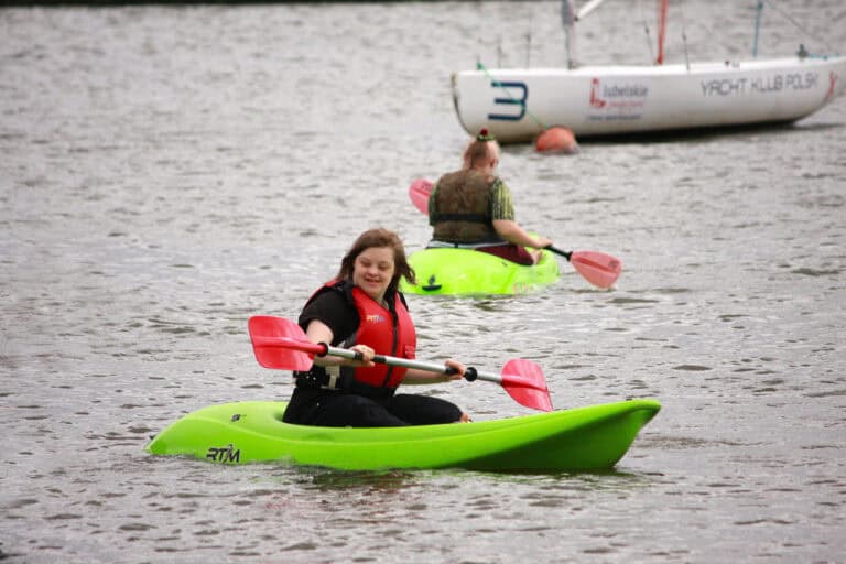 Na pierwszym planie w jaskrawo zielonym kajaku płynie młoda uśmiechnięta kobieta. Trzyma w rękach wiosło. Na drugim planie z prawej kobieta w kajaku, za nią żaglówka