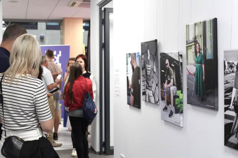 Od lewej grupa zwiedzających wystawę. Z prawej fotografie rozmieszczone asymetrycznie na tle jasnej ściany