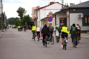 Kilka kilkunastoosobowych grup rowerzystów pokazanych z perspektywy tylnej. Rowerzyści poruszają się drogą, przy której ciągną się zabudowania mieszkalne i sklepy. W tle jasne niebo i zieleń drzew