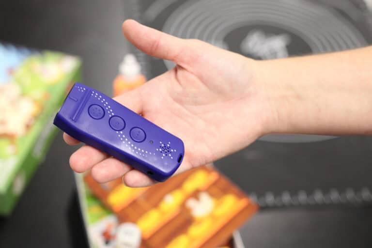 Dłoń, na której leży niebieski udźwiękowiony identyfikator (tester) kolorów dla osób niewidomych. Urządzenie rozpoznaje barwy i wypowiada je głosem