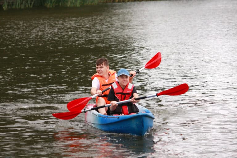 Na pierwszym planie w niebieskim kajaku płyną dwaj młodzieńcy w pomarańczowych kapokach. Trzymają w dłoniach wiosła