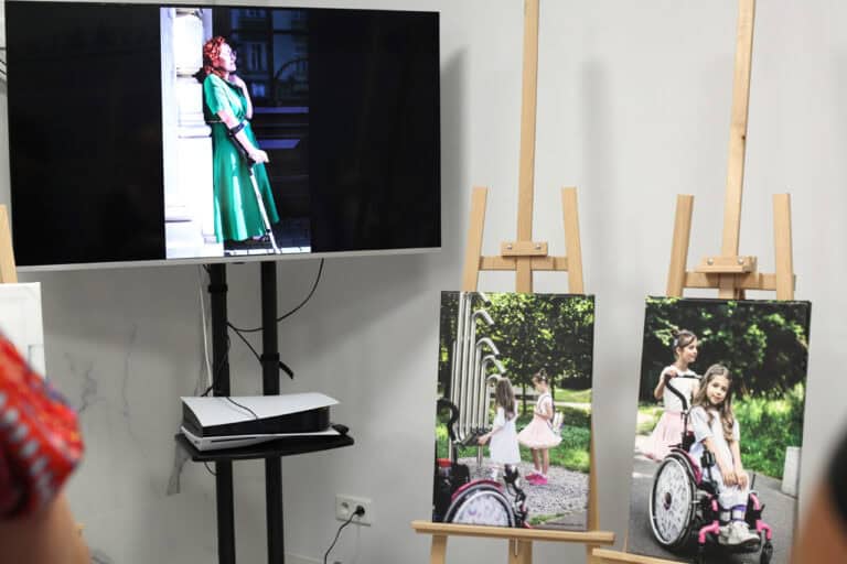 Od lewej: na monitorze wyświetlona fotografia kobiety w zielonej sukience. Kobieta wspiera się na kuli. Obok monitora stoją dwie sztalugi, na których wyeksponowano dwie fotografie małych dziewczynek