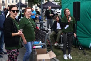 Na pierwszym planie trzy kobiety w zielonych koszulkach. Kobieta z lewej ma na sobie dodatkowo czarną bluzę i ciemne okulary. Kobieta z prawej uśmiechając się trzyma przy ustach mikrofon. W tle zgromadzeni ludzie, czarne parasole stoisk i zabudowania mieszkalne